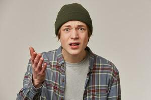 Portrait of confused, displeased male with blond hair. Wearing checkered shirt and beanie. People and emotion concept. Points palm at you, asking. Watching at the camera isolated over grey background photo