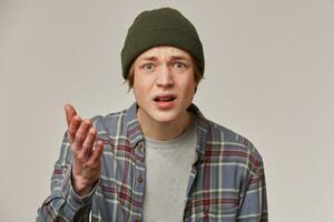 Portrait of confused, displeased male with blond hair. Wearing checkered shirt and beanie. People and emotion concept. Points palm at you, asking. Watching at the camera isolated over grey background photo