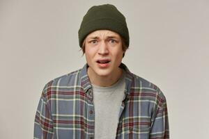 Discontent looking male, confused guy with blond hair. Wearing checkered shirt and beanie. People and emotion concept. Watching with disgust at the camera isolated over grey background photo