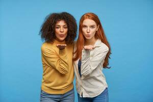 Indoor shot of positive charming young pretty women raising their palms and folding lips while blowing air kiss at camera, standing against blue background photo