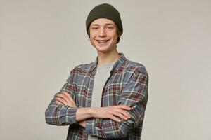 Cool looking male, handsome guy with blond hair. Wearing checkered shirt and beanie. Has braces. Holds arms crossed. People and emotion concept. Watching at the camera isolated over grey background photo