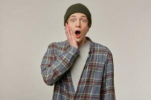 Portrait of shocked, scared male with blond hair. Wearing checkered shirt and beanie. People and emotion concept. Touching his face, opens mouth. Watching at the camera isolated over grey background photo