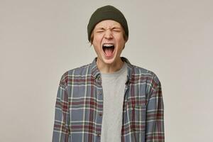 Teenage guy, unhappy looking man with blond hair. Wearing checkered shirt and beanie. Has braces. Keeps his eyes closed and yell. People and emotion concept. Stand isolated over grey background photo