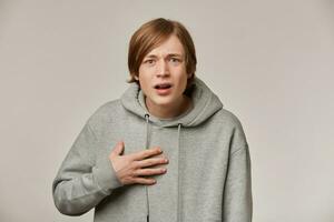 Portrait of confused, bewildered male with blond hair. Wearing grey hoodie. People and emotion concept. Pointing at himself with unsure. Watching at the camera isolated over grey background photo