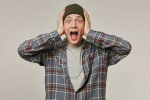 Portrait of terrified, adult male with blond hair. Wearing checkered shirt and beanie, has braces. Holding his head and scream. Emotion concept. Watching at the camera isolated over grey background photo