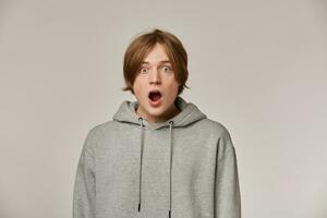 Portrait of shocked, amazed male with blond hair. Wearing grey hoodie. People and emotion concept. Stunned from what he sees. Watching surprised at the camera isolated over grey background photo