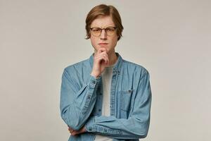 Cool looking male, focused guy with blond hair. Wearing denim shirt and glasses. Touching his chin and frowns. Emotion concept. Watching thoughtfully at the camera isolated over grey background photo