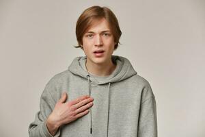 Portrait of confused, adult male with blond hair. Wearing grey hoodie. People and emotion concept. Pointing at himself and asking. Watching frowning at the camera isolated over grey background photo