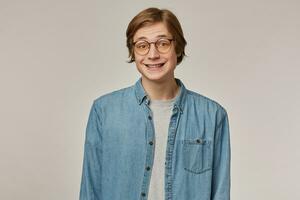 Adolescente chico, contento mirando hombre con rubio cabello. vistiendo azul mezclilla camisa, lentes y tiene tirantes. se encoge de hombros y sonriente. emoción concepto. acecho a el cámara aislado terminado gris antecedentes foto