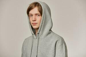 Cool looking male, handsome guy with blond hair. Wearing grey hoodie and puts hood on. People and emotion concept. Watching serious at the camera isolated over grey background photo