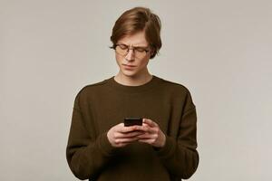 Serious looking male, handsome guy with blond hair. Wearing brown sweater and glasses. Holding a smartphone and frowns from what he read. Stand isolated over grey background photo