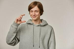 Teenage guy, tricky looking man with blond hair. Wearing grey hoodie. People and emotion concept. Showing a little size and smiles slyly. Watching at the camera isolated over grey background photo