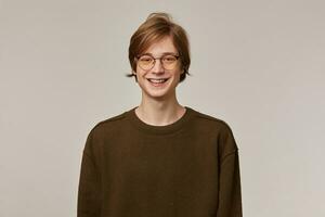 Cool looking male, cheerful guy with blond hair. Wearing brown sweater and glasses. Has braces. People and emotion concept. Watching with big smile at the camera isolated over grey background photo
