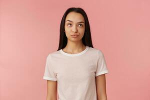 Teenage girl, annoyed looking woman with dark long hair. Wearing white t-shirt. People and emotion concept. Rolls her eyes and pursed lips. Watching up at copy space, isolated over pink background photo