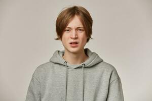 Irritated looking male, displeased guy with blond hair. Wearing grey hoodie. Has braces. People and emotion concept. Frowns his face and watching at the camera isolated over grey background photo