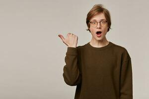 Shocked looking male, handsome guy with blond hair. Wearing brown sweater and glasses. Watching at the camera and pointing thumb to the left at copy space, isolated over grey background photo