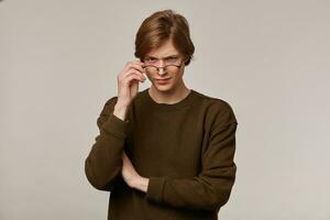 Serious teenage guy, unsure man with blond hair. Wearing brown sweater and glasses. Touching his glasses. People and emotion concept. Watching suspecting at the camera isolated over grey background photo