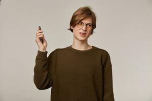 Frowning guy, unhappy looking man with blond hair. Wearing brown sweater and glasses. Has braces. Keeps phone away from ear. Annoying conversation. Stand isolated over grey background photo