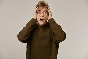 Teenage guy, terrified looking man with blond hair. Wearing brown sweater, glasses and has braces. Holds his head in fear. Emotion concept. Watching at the camera isolated over grey background photo