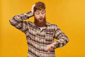 Young male student, with red hair and big beard, looks at his watches, keeps his hand on head and think about that he is late at work. Isolated over yellow background, photo