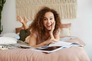 retrato de joven africano americano sorprendido mujer con Rizado cabello, mentiras en el cama y lee un nuevo revista, toques mejilla, en general sonrisas y disfrutar frio Noticias en revista, gasta gratis hora a hogar. foto