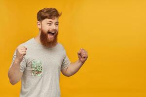 Indoor shot of young ginger bearded student, wears grey blank t-shirt, looks aside with happy facial expression, raised his fist while celebrate goal of his favorite football team. photo