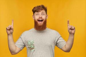 Indoor shot of young bearded male, wears grey t-shirt, pointing with both hands at copy space above him, smiling with amazed facial expression. Isolated over yellow background. photo