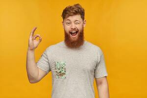 young male student, wears grey t-shirt, with red hair and beard, shows ok sign, smiles broadly and winks. Isolated over yellow background photo