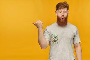 indoor shot of amazed, positive young bearded male with red hair, pointing with a finger left at copy space with amazed facial expression. Isolated over yellow background photo