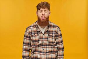 indoor shot of sad, resentful young bearded male, wears casual shirt, keeps his eye closed and shows his tongue to girlfriend. Isolated over yellow background photo