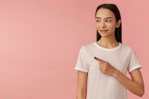 Cute looking woman, beautiful girl with long black hair. Wearing white t-shirt. People concept. Watching and pointing with index finger to the left at copy space, isolated over pastel pink background photo