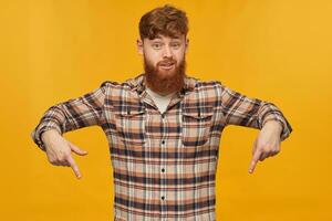 Young bearded male, wears casual shirt, with big beard and red hair, indicates with both hands down, isolated over yellow background photo