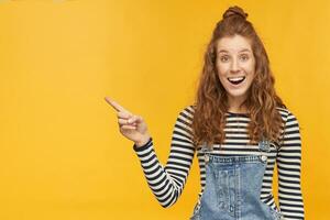 indoor portrait of young ginger curly female posing over yellow bright background points with a finger aside at copy space with shocked facial expression and smiles broadly photo