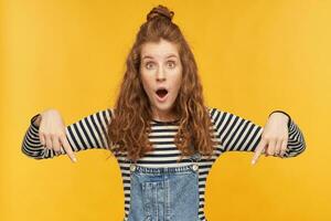 Indoor shot of young female with a red long hair, indicates with a finger downward at your copy space with shocked, amazed facial expression. isolated over yellow background. photo