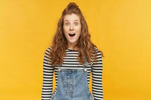 indoor portrait of amazed young ginger girl, wears denim overalls and stripped shirt, starring into camera with widely opened mouth and eyes. shocked, amazed facial expression. photo