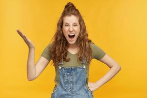 Negative, mad ginger female, wears blue denim overalls and green t-shirt, screaming and raised her hand with negative facial expression. isolated over yellow background. photo