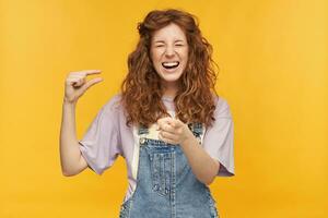 young ginger female with a long wavy hair shows something small with one hand, pointing at you with another an laughing, isolated over yellow background photo