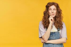 Portrait of pensive young ginger female with long wavy hair, looks aside at copy space thoughtfully, toughing her chin and thinking about future. isolated over yellow background. photo