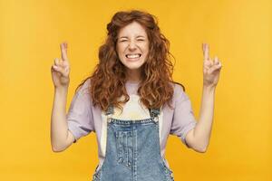 indoor shot of young female student wears blue denim overalls and purple t-shirt, crossed her fingers in praying position, waiting for a good exams result. isolated over yellow background. photo