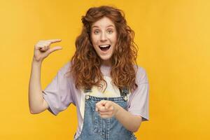 young ginger female with a long wavy hair shows something small with one hand, pointing at you with another an laughing, isolated over yellow background photo