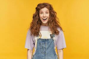 young ginger female posing against yellow studio background with amazed, shocked facial expression, smiles broadly and keep eyes widely opened. Isolated photo