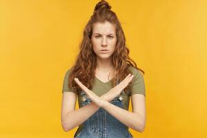 indoor shot of displease, unhappy young ginger female, shows no sign with both hands, looking into camera with negative facial expression. isolated over yellow background. photo