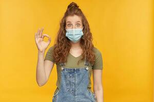 indoor studio shot of young ginger female with long red curly hair shows ok sign with positive, happy facial expression, wears face mask, isolated over yellow background photo