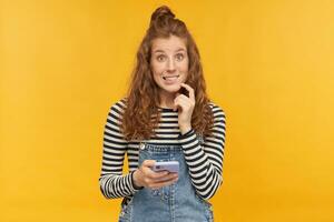 indoor studio shot of young ginger female with long red curly hair biting her lip, holding phone with ashamed guilty facial expression, isolated over yellow background photo