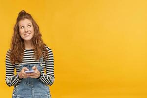 indoor studio shot of young ginger female with long red curly hair looks aside at copy space with ashamed, surprised facial expression, isolated over yellow background photo