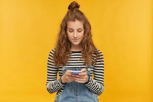 indoor studio shot of young ginger female with long red curly hair keep calm while typing message on her phone isolated over yellow background photo