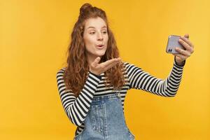 Indoor shot of young female talking via phone with her boyfriend, feels pleased and happy, sending air kiss, isolated over yellow background. photo