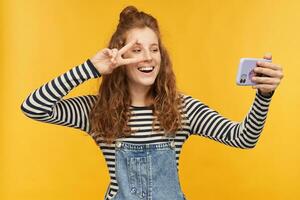 indoor portrait of young happy female, wears stripped shirt and denim overalls shows v sign while have video chat with her boyfriend while isolation. photo