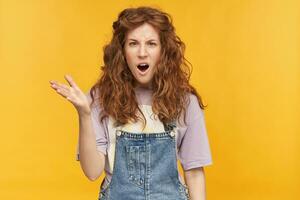 young negative female, wears blue overalls and purple t-shirt, raised her hand with unhappy, mad facial expression, keeps hey mouth widely opened. Isolated over yellow background photo