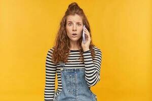 indoor studio shot of young ginger female with long red curly hair talking on the phone with confused, guilty facial expression, isolated over yellow background photo
